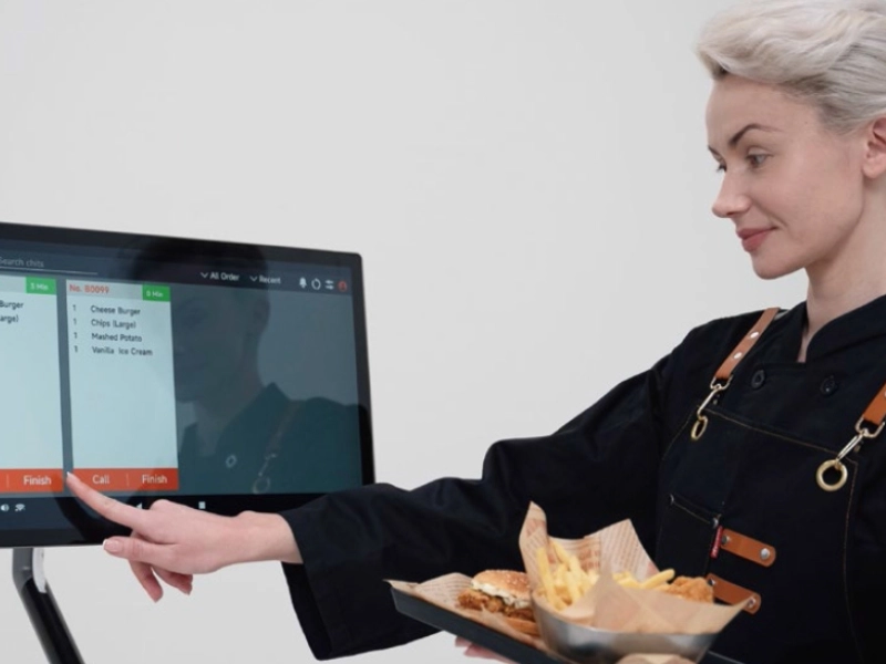 Servicekraft im Restaurant tippt auf ein Touchscreen-Kassensystem und trägt ein Tablett mit Burger und Pommes.